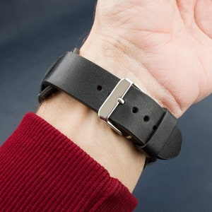 Puede incluir: Una correa de reloj de cuero negro con una hebilla plateada. La correa tiene dos agujeros para ajustar el tama&ntilde;o.