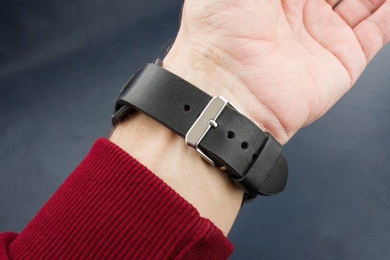 Puede incluir: Una correa de reloj de cuero negro con una hebilla plateada. La correa tiene dos agujeros para ajustar el tama&ntilde;o.