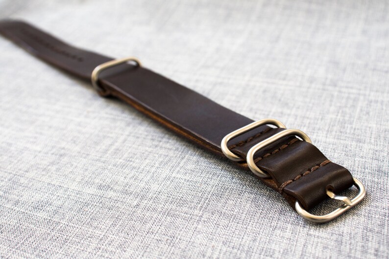 Puede incluir: Una correa de reloj de cuero marr&oacute;n con anillos y hebilla de metal plateado.