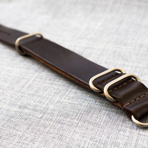 Puede incluir: Una correa de reloj de cuero marr&oacute;n con anillos y hebilla de metal plateado.