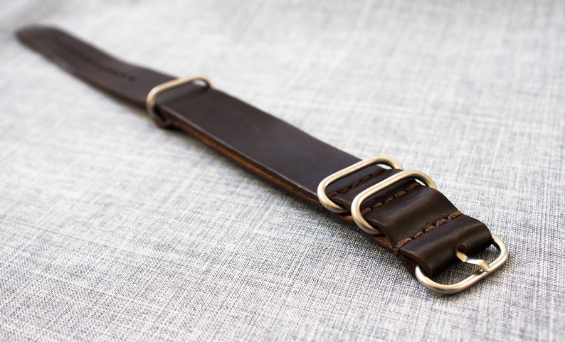 Puede incluir: Una correa de reloj de cuero marr&oacute;n con anillos y hebilla de metal plateado.