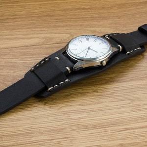 Puede incluir: Una correa de reloj de cuero negro con costuras blancas y una esfera de reloj plateada con n&uacute;meros romanos blancos.
