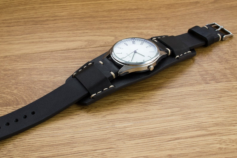 Puede incluir: Una correa de reloj de cuero negro con costuras blancas y una esfera de reloj plateada con n&uacute;meros romanos blancos.