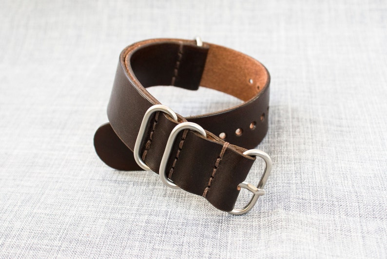 Puede incluir: Una correa de reloj de cuero marr&oacute;n con herrajes y costuras plateadas. La correa tiene varios agujeros para ajustar el ajuste.