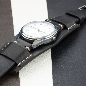Puede incluir: Una correa de reloj de cuero negro con costuras blancas. La correa est&aacute; envuelta alrededor de un reloj plateado con una esfera blanca y agujas negras.