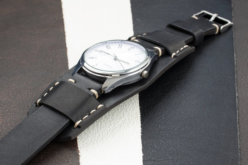 Puede incluir: Una correa de reloj de cuero negro con costuras blancas. La correa est&aacute; envuelta alrededor de un reloj plateado con una esfera blanca y agujas negras.