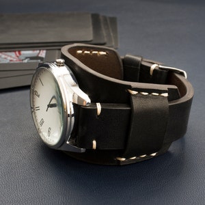 Puede incluir: Un reloj de pulsera plateado con una esfera blanca y marcadores de hora romanos negros en una correa de reloj de brazalete de cuero negro.