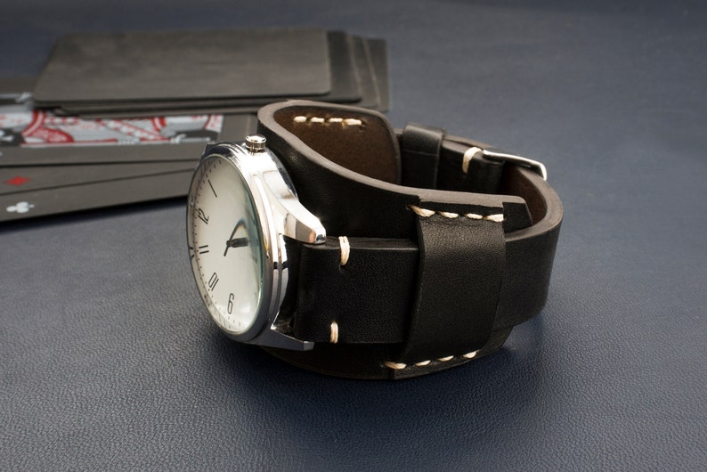 Puede incluir: Un reloj de pulsera plateado con una esfera blanca y marcadores de hora romanos negros en una correa de reloj de brazalete de cuero negro.