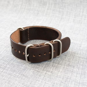 Puede incluir: Una correa de reloj de cuero marr&oacute;n con hebilla y trabillas plateadas.