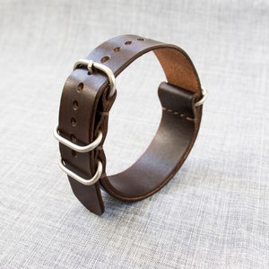 Puede incluir: Una correa de reloj de cuero marr&oacute;n con hebilla y trabillas de metal plateado.