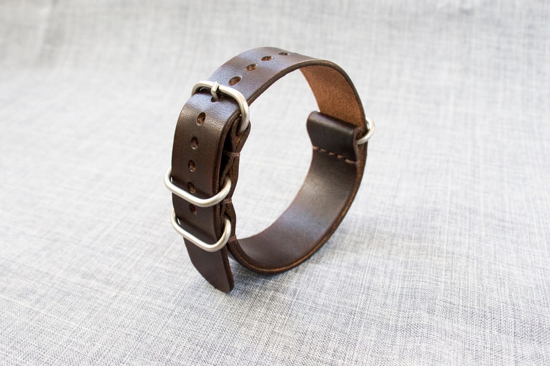 Puede incluir: Una correa de reloj de cuero marr&oacute;n con hebilla y trabillas de metal plateado.