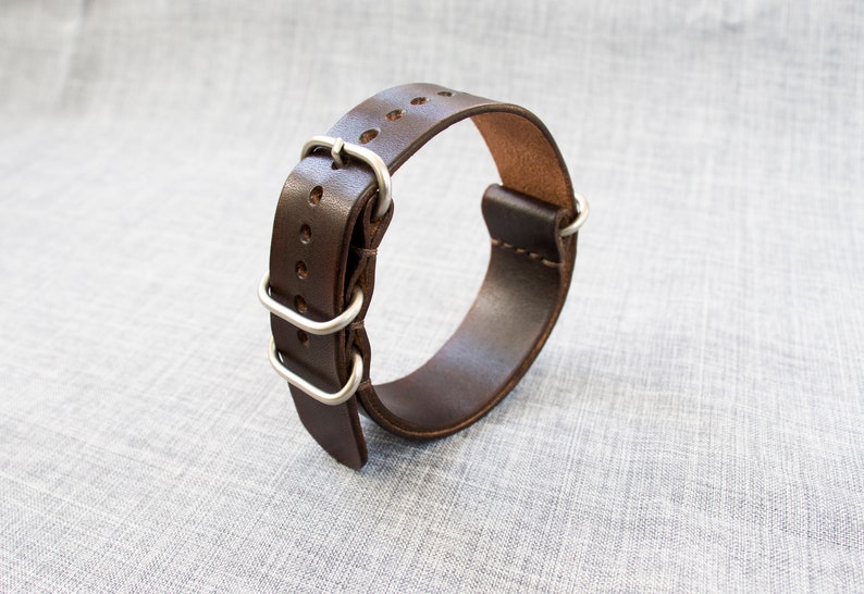 Puede incluir: Una correa de reloj de cuero marr&oacute;n con hebilla y trabillas plateadas.