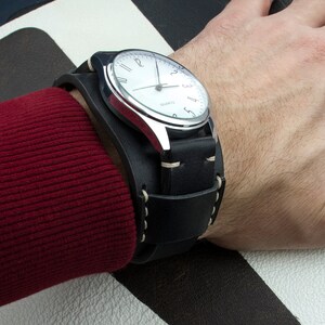 Puede incluir: Un reloj de pulsera plateado con una esfera blanca y una correa de brazalete de cuero negro. El reloj tiene marcadores de hora en n&uacute;meros romanos y una aguja de segundos blanca.