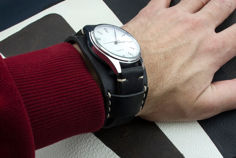 Puede incluir: Un reloj de pulsera plateado con una esfera blanca y una correa de brazalete de cuero negro. El reloj tiene marcadores de hora en n&uacute;meros romanos y una aguja de segundos blanca.