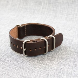 Puede incluir: Una correa de reloj de cuero marr&oacute;n con hebilla y trabillas plateadas.