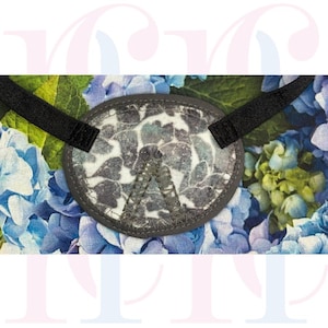 Può includere: Una benda sull'occhio con un motivo floreale nei toni del blu, grigio e bianco, su uno sfondo di fiori di ortensia blu. La benda ha un bordo grigio e cinturini neri per un uso sicuro.