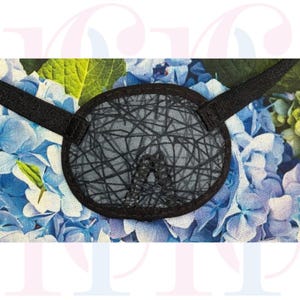Könnte beinhalten: Eine Augenklappe mit schwarzem Rand und grauem Zentrum mit einem netzartigen Design. Die Augenklappe hat schwarze Riemen und ist vor einem Hintergrund aus blauen und grünen Hortensienblüten angeordnet.