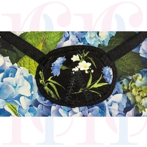 May include: Black eye patch with a floral design featuring blue and white flowers and green leaves. The eye patch has a black elastic strap and is set against a background of blue and green floral fabric.
