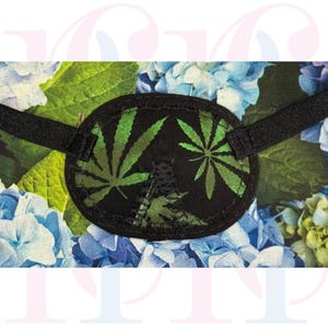 May include: Black eye patch featuring a green cannabis leaf design. The oval patch has a black border and an adjustable black strap. The background shows blue and green floral patterns.