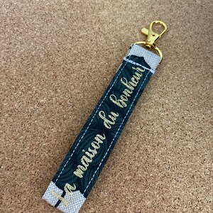 K&ouml;nnte beinhalten: Ein goldener Schl&uuml;sselanh&auml;nger mit einem wei&szlig;en und gr&uuml;nen Stoffband. Das Band tr&auml;gt den Text "La maison du bonheur" in goldenen Buchstaben.
