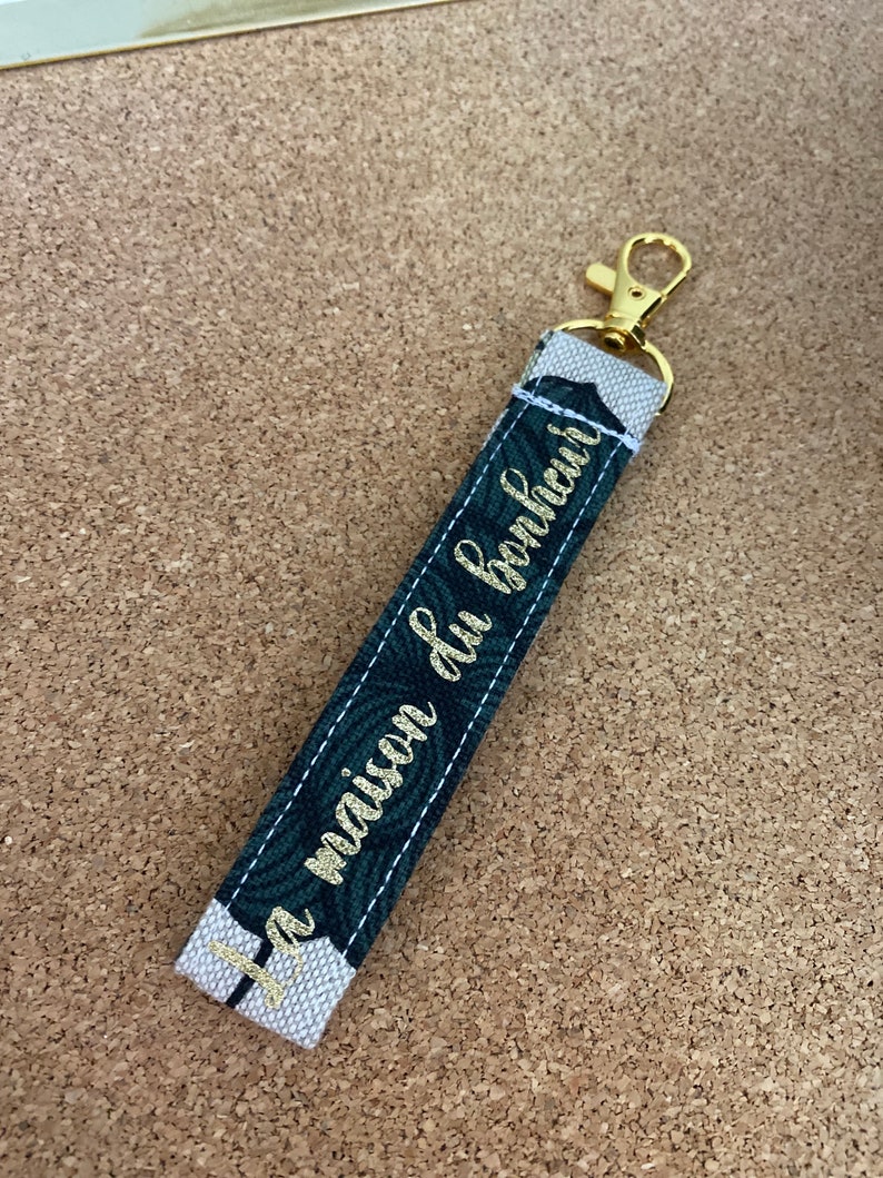 K&ouml;nnte beinhalten: Ein goldener Schl&uuml;sselanh&auml;nger mit einem wei&szlig;en und gr&uuml;nen Stoffband. Das Band tr&auml;gt den Text "La maison du bonheur" in goldenen Buchstaben.