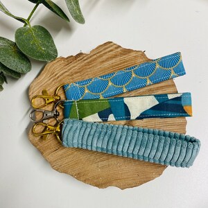 K&ouml;nnte beinhalten: Drei Schl&uuml;sselanh&auml;nger mit verschiedenen Stoffmustern. Der obere Schl&uuml;sselanh&auml;nger ist blau mit einem goldenen geometrischen Muster. Der mittlere Schl&uuml;sselanh&auml;nger ist gr&uuml;n, wei&szlig; und blau mit einem geometrischen Muster. Der untere Schl&uuml;sselanh&auml;nger ist aus einfarbigem blauem Cord.
