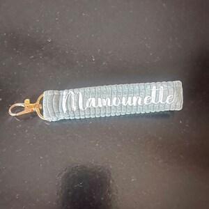 K&ouml;nnte beinhalten: Grauer Stoffschl&uuml;sselanh&auml;nger mit einem goldenen Metallring und dem in Wei&szlig; gestickten Wort "Mamounette".