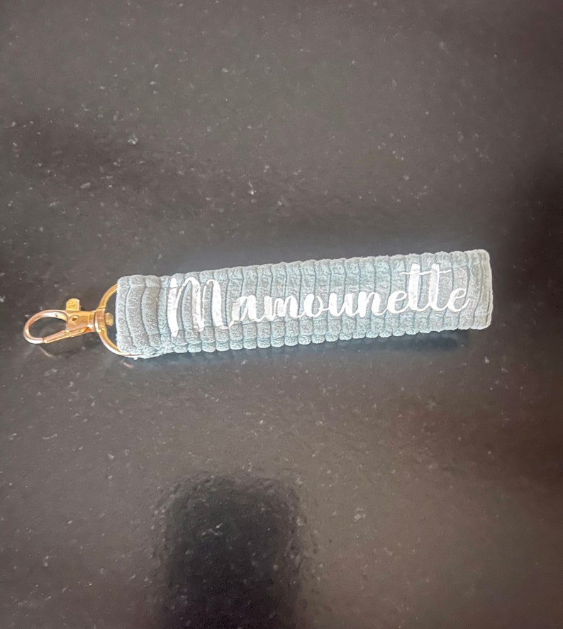 K&ouml;nnte beinhalten: Grauer Stoffschl&uuml;sselanh&auml;nger mit einem goldenen Metallring und dem in Wei&szlig; gestickten Wort "Mamounette".