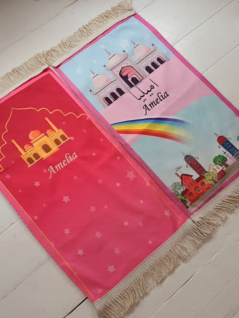 May include: Two pink prayer rugs with fringe trim. One rug features a mosque with a rainbow and the name Amelia. The other rug features a mosque and stars with the name Amelia.