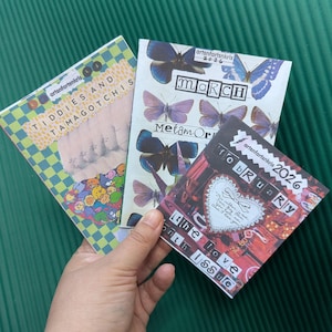 May include: A collection of three zines with colorful covers. One zine features the text "Tiddies and Tamagotchis" with a checkered background. Another displays butterflies and the text "March". The third zine says "February 2026" and "The Love Month Issue".