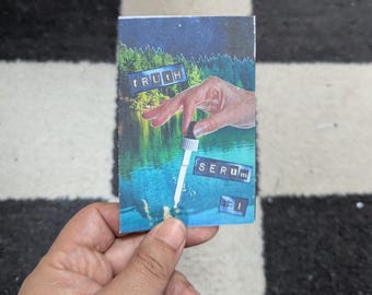 May include: A small, rectangular card featuring a collage with a serene lake scene and the words "TRUTH SERUM #1". A hand is depicted holding a dropper, suggesting a skincare or cosmetic product.