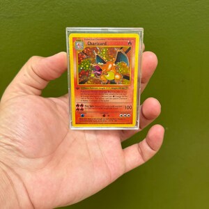 May include: A Charizard Pokémon trading card in a protective case. The card features a vibrant orange and yellow design with a dragon-like creature. The card has the number 100 and the name "Charizard" printed on it.