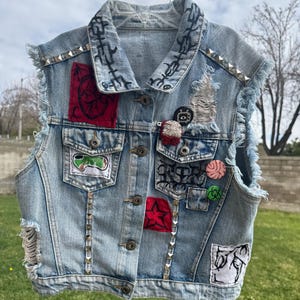 May include: Light blue denim vest with patches, studs, and distressed details. The vest features various patches with embroidered designs, including a red heart, a green graphic, and a white and black illustration. Silver studs line the edges.