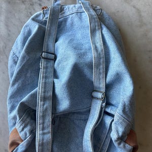 May include: A light blue denim backpack with adjustable straps and a top handle. The backpack features brown leather accents on the bottom corners and black metal hardware. The bag is designed for carrying items.