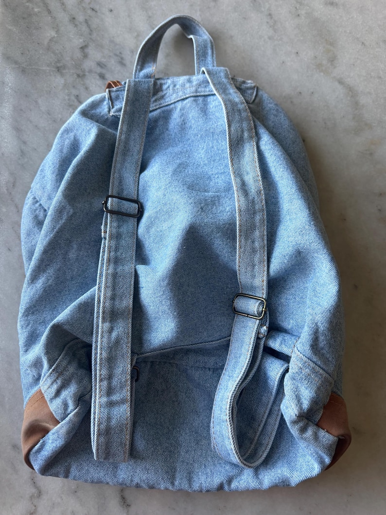 May include: A light blue denim backpack with adjustable straps and a top handle. The backpack features brown leather accents on the bottom corners and black metal hardware. The bag is designed for carrying items.