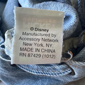 May include: Close-up of a white tag with black text on a light blue denim garment. The tag reads "&copy; Disney Manufactured by Accessory Network New York, NY. MADE IN CHINA RN 87429 (1012)".