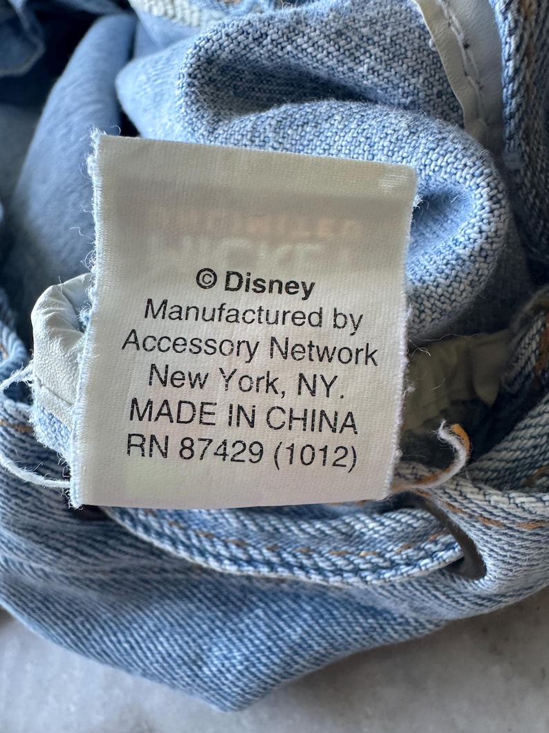 May include: Close-up of a white tag with black text on a light blue denim garment. The tag reads "&copy; Disney Manufactured by Accessory Network New York, NY. MADE IN CHINA RN 87429 (1012)".