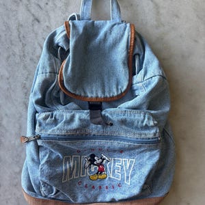 May include: A light blue denim backpack with brown leather trim. The front pocket features an embroidered Mickey Mouse graphic with the word "MICKEY" in white block letters. The backpack has a top handle and adjustable straps.