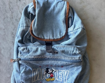 May include: A light blue denim backpack with brown leather trim. The front pocket features an embroidered Mickey Mouse graphic with the word "MICKEY" in white block letters. The backpack has a top handle and adjustable straps.
