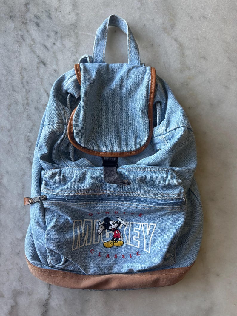 May include: A light blue denim backpack with brown leather trim. The front pocket features an embroidered Mickey Mouse graphic with the word "MICKEY" in white block letters. The backpack has a top handle and adjustable straps.