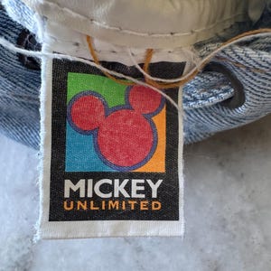 May include: Close-up of a denim garment with a "Mickey Unlimited" tag. The tag features a red Mickey Mouse head on a black background with colorful accents. The garment's waistband and stitching are visible.