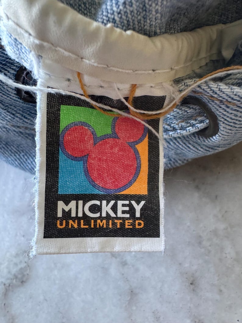 May include: Close-up of a denim garment with a "Mickey Unlimited" tag. The tag features a red Mickey Mouse head on a black background with colorful accents. The garment's waistband and stitching are visible.
