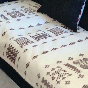 May include: Cream-coloured textile featuring a repeating geometric pattern in brown. The fabric is draped over a dark surface, with a black cushion in the upper right corner. The pattern includes diamond and rectangular shapes.