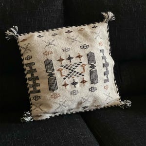Può includere: Cuscino quadrato color crema con un motivo geometrico nero, marrone e grigio. Il cuscino ha un bordo intrecciato bianco e nero e nappe ad ogni angolo. Il cuscino è su una superficie nera.