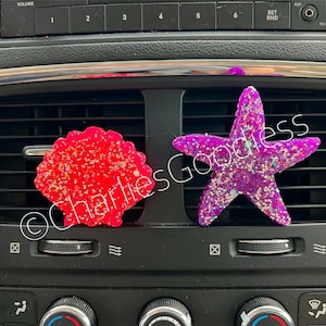 May include: Two car air fresheners, one shaped like a seashell and the other like a starfish. The seashell is red with glitter and the starfish is purple with glitter.