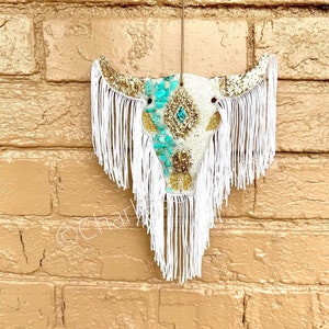 May include: White cow skull with gold glitter and turquoise accents, decorated with white fringe.