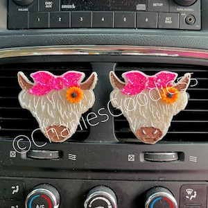 May include: Two white fuzzy cow car air fresheners with pink bows and orange flowers. The air fresheners are hanging from the air vent in a car.