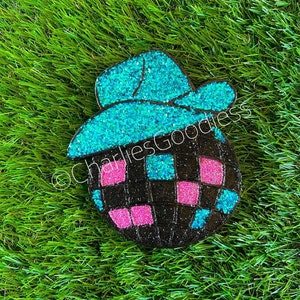 May include: A glittery, black disco ball ornament with pink and blue squares, topped with a turquoise cowboy hat. The ornament is set against a backdrop of green grass, with the text "@CharliesGoodless" visible.
