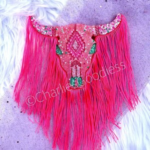 May include: Pink and green glitter skull with fringe. The skull has a geometric pattern and is surrounded by pink fringe.