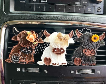 May include: Three car air fresheners shaped like Highland cows. The cows are brown, white, and black with glitter accents. Each cow has a different colored flower on its head.